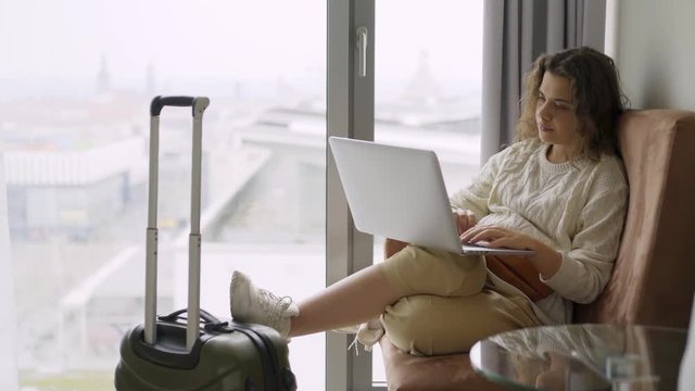 Young woman arrived modern hotel suite. Side view girl sitting in armchair near full length window with laptop. She relaxing after journey, working on computer and enjoying view above city