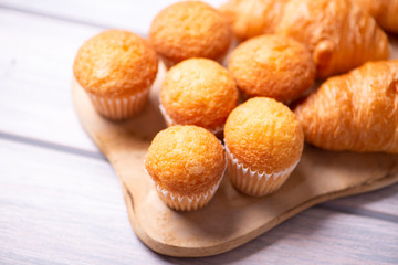 Close-up of cupcakes and croissants prepared for breakfast on wooden board on white wooden table. Copy space to the left of the image.