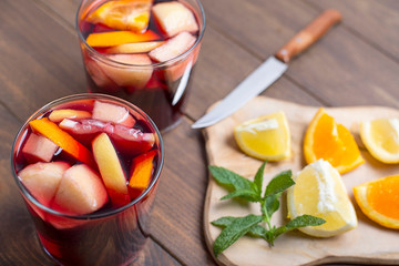 Two glasses with sangria and fruits next to table with lemon and orange cut on top next to knife....