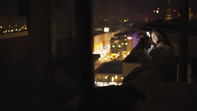 Woman relaxing in modern hotel suite against big window at night time. Side view sitting girl in bathrobe focused on smartphone and drinking tea in silhouette at background view city lights in bokeh