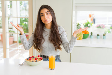 Young woman eating healthy breakfast in the morning clueless and confused expression with arms and...