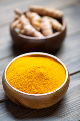 Vertical shoot of bowl with curcumin powder in the foreground next to another bowl with turmeric root on brown wooden table. Spice for health and ingredient of Indian cuisine.
