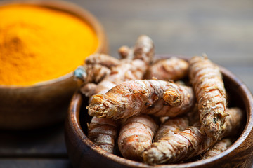 Bowl with turmeric root in the foreground next to bowl with turmeric powder on brown wooden table. Spice for health and ingredient of Indian cuisine.