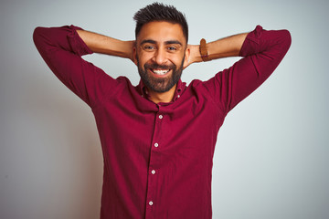 Young indian man wearing red elegant shirt standing over isolated grey background relaxing and stretching, arms and hands behind head and neck smiling happy