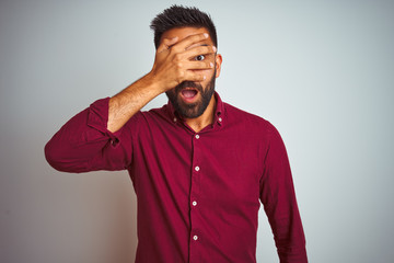 Young indian man wearing red elegant shirt standing over isolated grey background peeking in shock covering face and eyes with hand, looking through fingers with embarrassed expression.