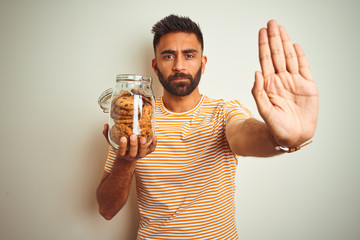 Young indian man holding jar of cookies standing over isolated white background with open hand...