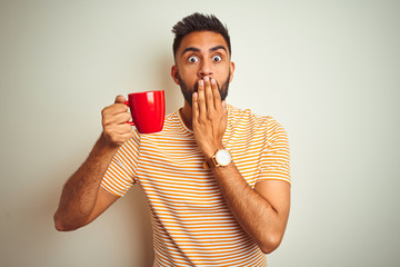 Young indian man drinking red cup of coffee standing over isolated white background cover mouth...