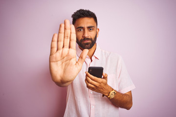 Young indian man using smartphone standing over isolated pink background with open hand doing stop...