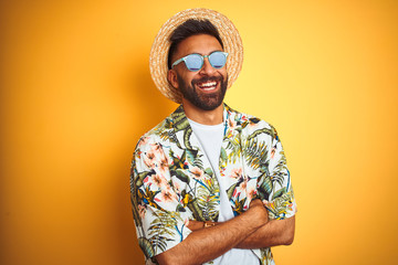 Indian man on vacation wearing floral shirt hat sunglasses over isolated yellow background happy...