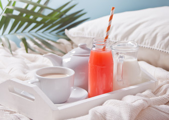 Summer tropical breakfast with cup of tea, teapot and refreshing exotic juice in a bottle with straw on a white tray under the palm leaf