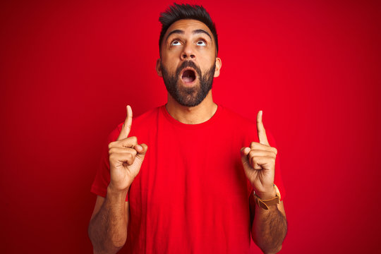 Young handsome indian man wearing t-shirt over isolated red background amazed and surprised looking up and pointing with fingers and raised arms.
