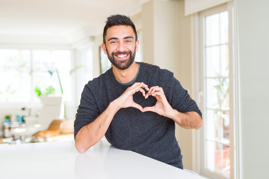 Handsome hispanic man wearing casual sweater at home smiling in love showing heart symbol and shape with hands. Romantic concept.