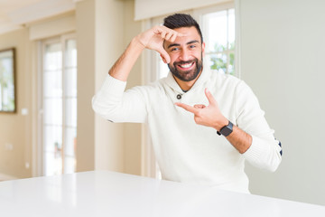 Fototapeta premium Handsome hispanic man wearing casual white sweater at home smiling making frame with hands and fingers with happy face. Creativity and photography concept.