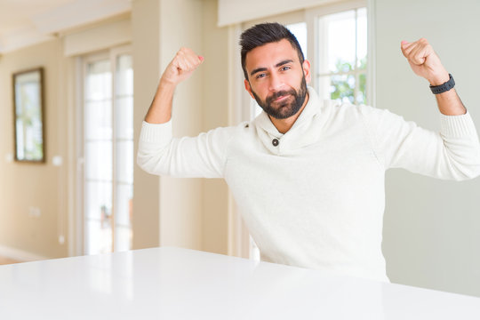 Handsome hispanic man wearing casual white sweater at home showing arms muscles smiling proud. Fitness concept.