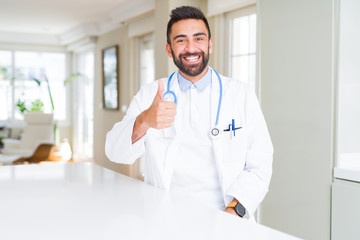 Handsome hispanic doctor man wearing stethoscope at the clinic doing happy thumbs up gesture with...