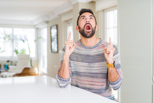 Handsome hispanic man wearing casual sweater at home amazed and surprised looking up and pointing with fingers and raised arms.
