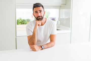 Handsome hispanic man casual white t-shirt at home with hand on chin thinking about question, pensive expression. Smiling with thoughtful face. Doubt concept.