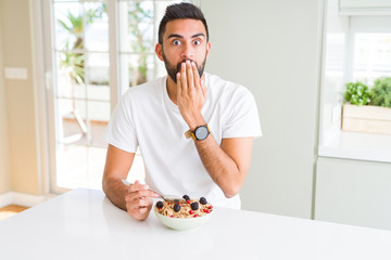 Handsome hispanic man eating healthy breakfast in the morning at home cover mouth with hand shocked with shame for mistake, expression of fear, scared in silence, secret concept