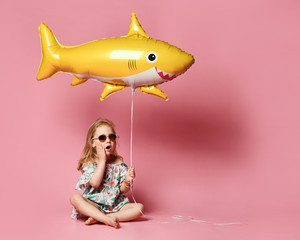 little girl, dressed in a summer dress, wears a balloon in the shape of a yellow shark-fish,...