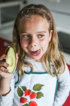Little girl with half of tasty fresh avocado looking away and licking lips while standing in kitchen