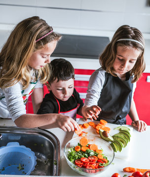 Little girls and boy cutting and peeling ripe vegetables while cooking healthy salad in kitchen together