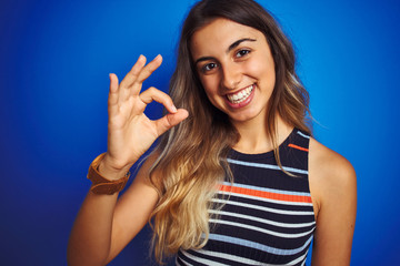 Young beautiful woman wearing stripes t-shirt over blue isolated background doing ok sign with fingers, excellent symbol