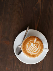 Overhead fresh brewed cappuccino coffee in white ceramic cup, saucer, spoon with beautiful handmade latte art on wooden table in cozy city cafe ready to drink