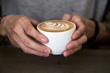 Male barista hands holding cup of fresh delicious cappuccino with latte art cream foam, brewed and made with love and take care concept, good service coffee shop
