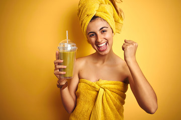 Young beautiful woman wearing a towel drinking detox juice over yellow isolated background...