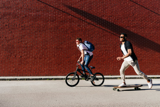 Side view of young black successful male friends in casual apparel and stylish sunglasses riding bike and skateboard
