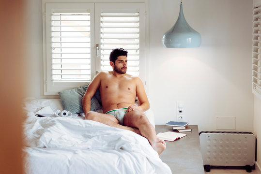 Young smiling successful sexy man with stylish hairstyle in grey briefs lying on bed at home and looking away