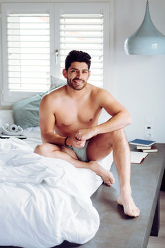Side view of young sexy thoughtful handsome male with stylish hairstyle in underwear sitting on bed and looking at camera