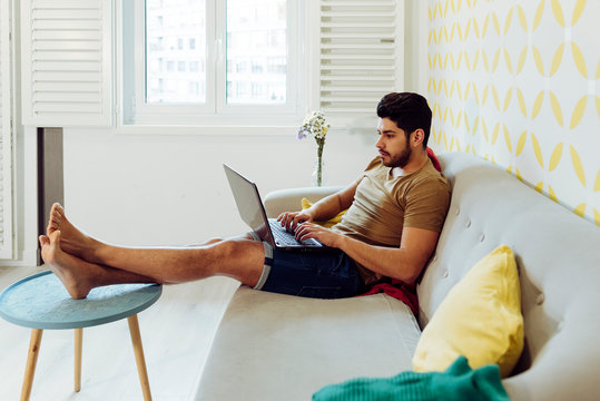 Side view of young thoughtful man in casual outfit with laptop sitting on couch near table with fresh flowers - Powered by Adobe