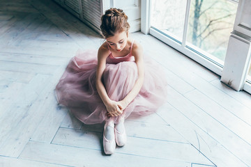Young classical ballet dancer girl in dance class. Beautiful graceful ballerina in pink tutu skirt puts on pointe shoes near large window in white light hall © Юлия Завалишина
