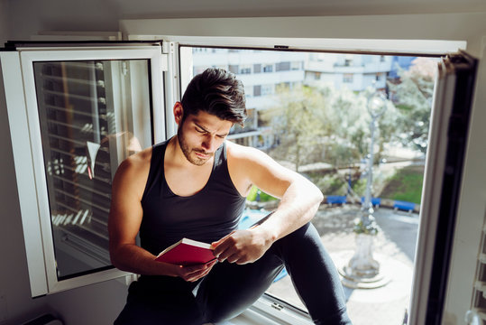 Side view of young serious handsome man in casual sleeveless shirt sitting on window and reading interesting book