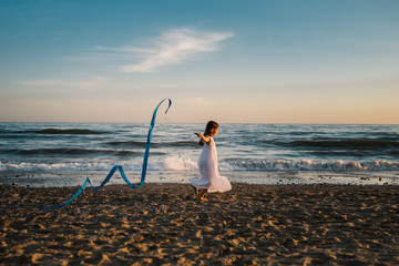 Little girl in white dress running along seashore and holding long blue band on background of evening sky