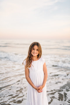 Portrait of charming little girl in white dress standing in water on sandy beach and looking at camera