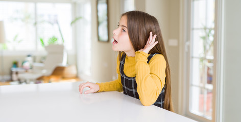 Beautiful young girl kid sitting on the table smiling with hand over ear listening an hearing to rumor or gossip. Deafness concept.