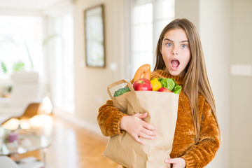 Beautiful young girl holding paper bag of fresh groceries scared in shock with a surprise face, afraid and excited with fear expression