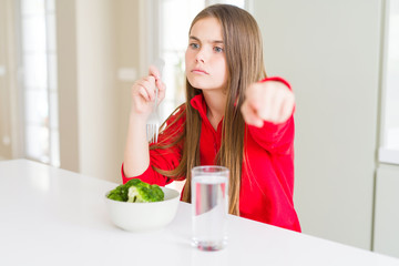 Beautiful young girl kid eating fresh broccoli and drinking water pointing with finger to the camera and to you, hand sign, positive and confident gesture from the front