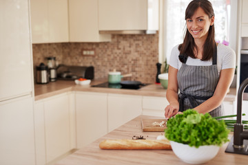 Smiling Caucasian female chef in apron chopping leek in kitchen and preparing dinner. Preparation of domestic food concept.