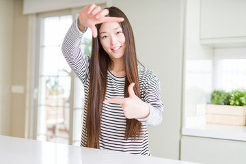 Beautiful Asian woman wearing stripes sweater smiling making frame with hands and fingers with happy face. Creativity and photography concept.