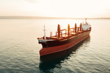 Obraz premium Aerial view of high-speed sea vessel for transportation of a cargo vessel at high speed is drifting near the seaport of the city at sunset. Ship on the background of blue sea water. Import, export
