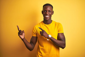 Young african american man wearing casual t-shirt standing over isolated yellow background smiling and looking at the camera pointing with two hands and fingers to the side.