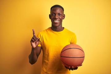 African american athlete man holding basketball ball standing over isolated yellow background...