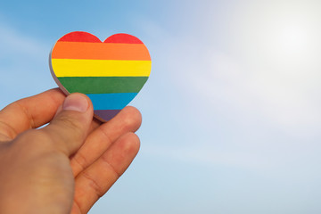 Man's hand  holds a heart painted like a LGBT gay flag on blue sky background. LGBT equal rights movement and gender equality concept. Gay pride homosexual concept.