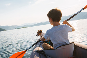 Happy boy with his dog Jack Russell Terrier paddling an inflatable kayak on the water mountain lake against the backdrop of beautiful orange sunset. Family sports vacation. Lens flare. Pet