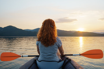 Fototapeta premium A happy red-haired woman looks into the distance on a calm lake surface in the mountains, in an inflatable kayak against the backdrop of an orange sunset. The sun's rays in her hair. Family tourism