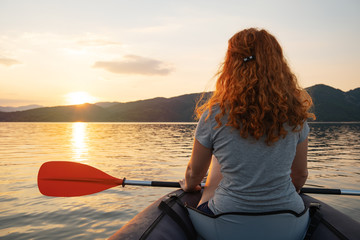 Fototapeta premium A happy red-haired woman looks into the distance on a calm lake surface in the mountains, in an inflatable kayak against the backdrop of an orange sunset. The sun's rays in her hair. Family tourism