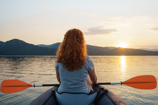 A happy red-haired woman looks into the distance on a calm lake surface in the mountains, in an inflatable kayak against the backdrop of an orange sunset. The sun's rays in her hair. Family tourism